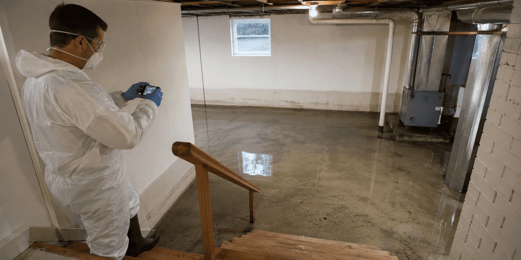A restoration technician documents shallow stormwater in a flooded basement from the dry stair landing.
